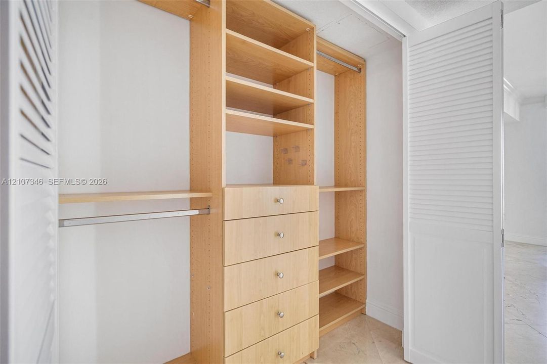 Walk-in closet featuring light wood-finish built-in shelving, a five-drawer dresser unit, stainless steel hanging rods, and white louvered doors