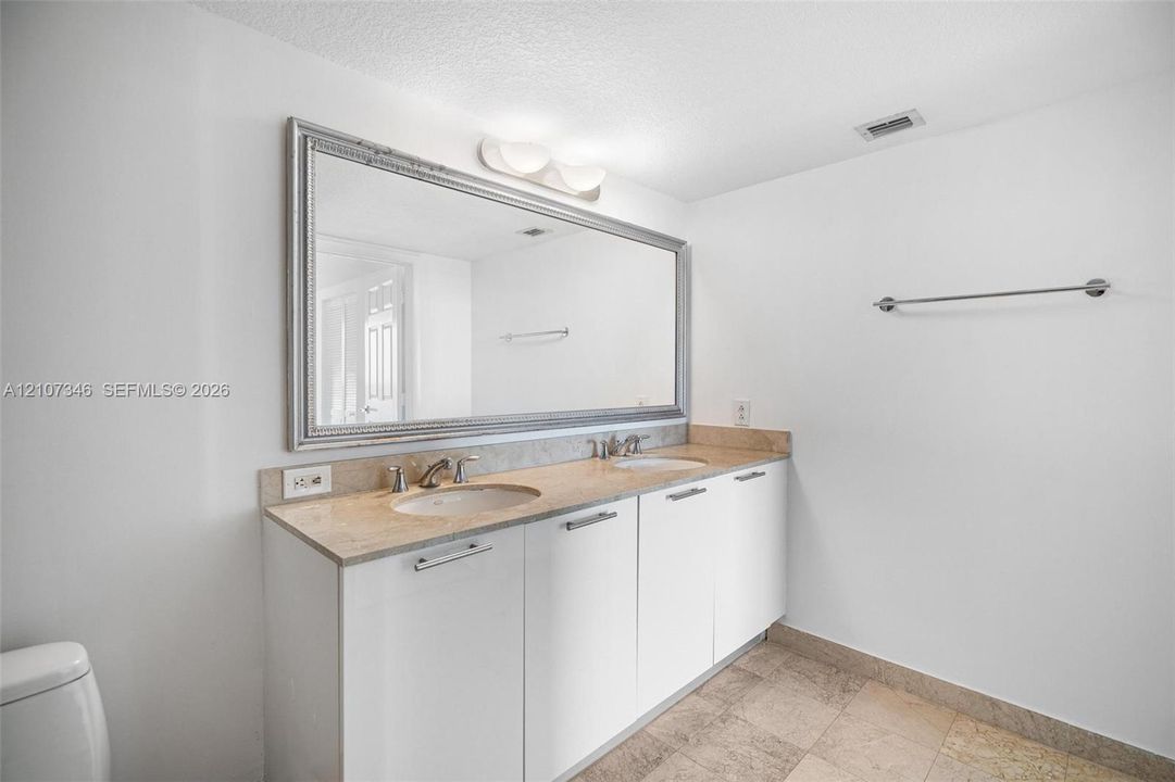 Dual-sink vanity with a stone countertop, white flat-panel cabinetry, large rectangular wall mirror, and integrated lighting