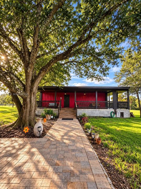 Exterior featuring a metal-sided structure with a covered front porch, concrete foundation, and a wide paver stone walkway beneath a large mature tree