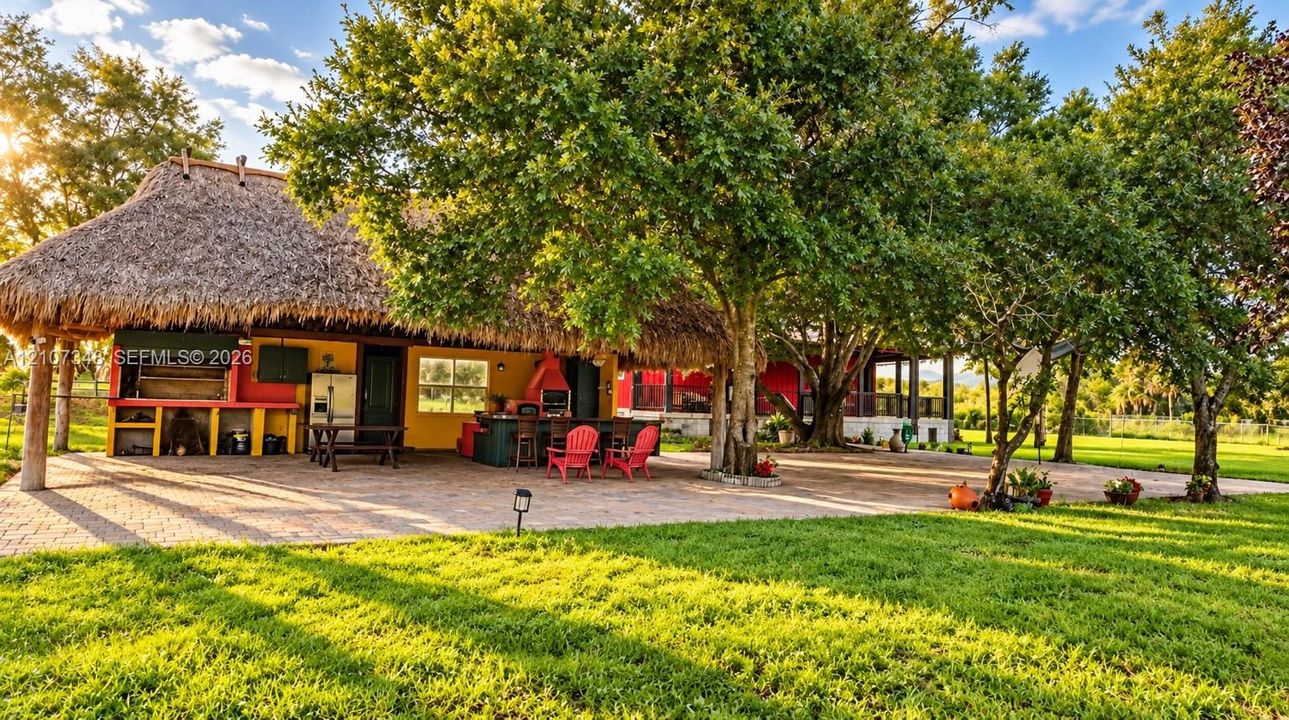 Outdoor living area featuring a thatch-roofed pavilion, stone-paver patio, built-in outdoor kitchen with a grill station, and mature landscaping