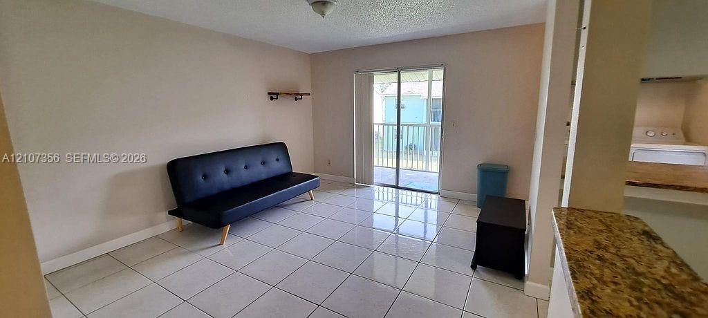 Looking from Hallway into Living/Dining Area and Balcony, Light-filled living area featuring ceramic tile flooring, sliding glass doors to a balcony, and a kitchen pass-through with stone-finish countertops