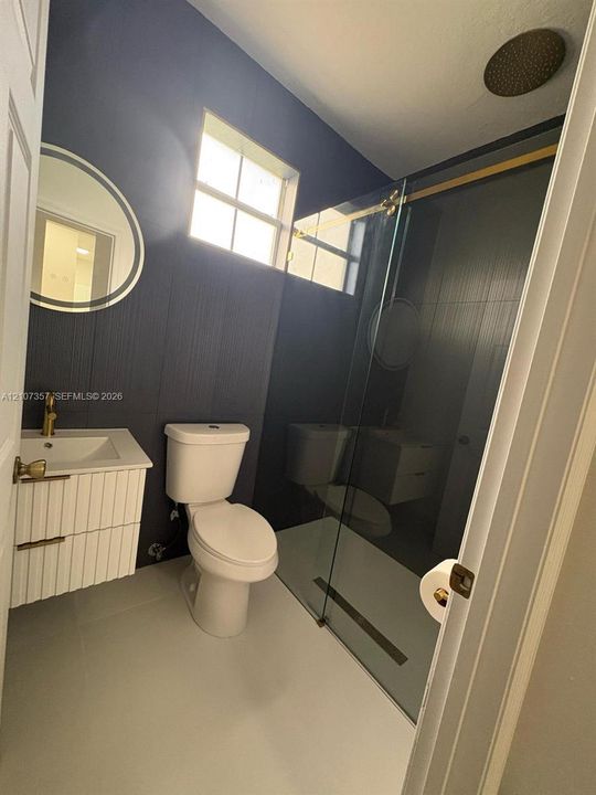 Wall-mounted white vanity with fluted cabinetry and gold-tone hardware, glass-enclosed walk-in shower with gold-tone fixtures, ceiling-mounted rainfall showerhead, and round LED-backlit mirror