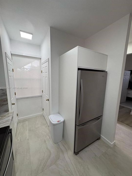 Stainless steel bottom-freezer refrigerator with integrated overhead cabinetry, large-format tile flooring, and flush-mount ceiling lighting