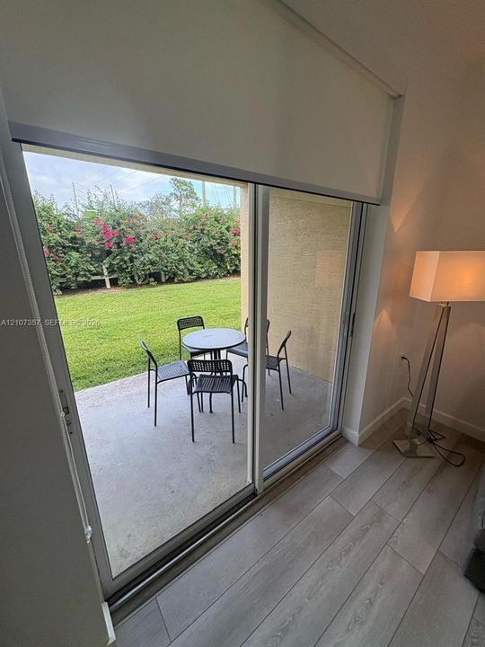 Large glass sliding doors, wood-finish flooring, and concrete patio leading to a manicured lawn with mature flowering shrubs