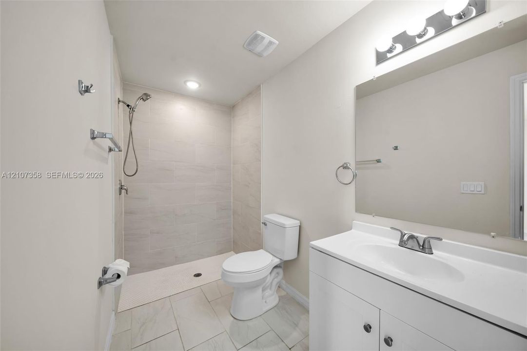 Full-size bathroom featuring a white vanity with an integrated basin, large wall-mounted mirror, light-bar fixture, walk-in shower with neutral tile surround, and tile flooring