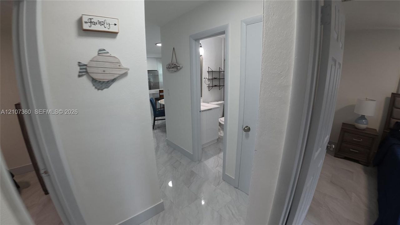 Interior hallway featuring large-format tile flooring, crisp white walls, and a recessed doorway providing entry to a bathroom equipped with a white vanity and stone-finish countertop