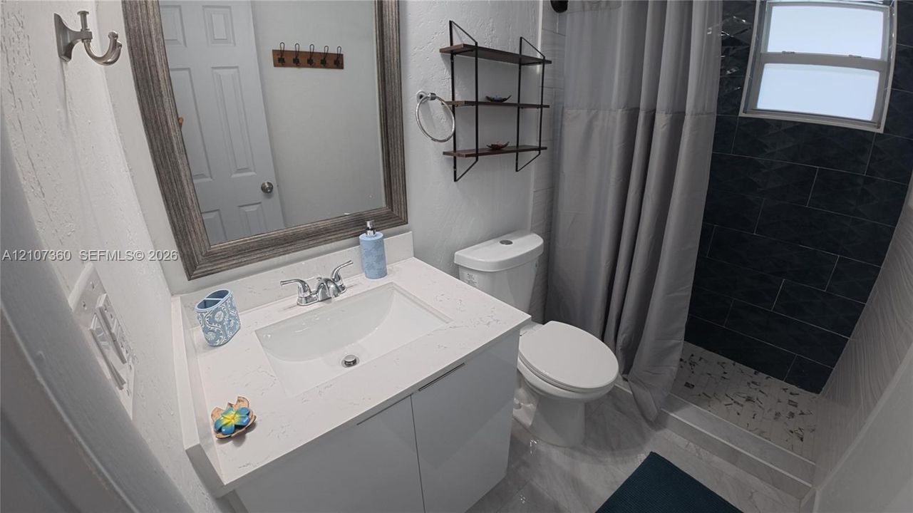 Bathroom featuring a vanity with a white composite countertop, a framed wall mirror, a shower with dark charcoal tile surround, and built-in metal shelving