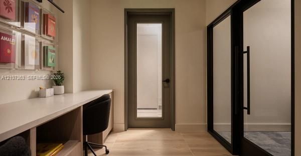 Built-in desk with extensive cabinetry, wood-finish flooring, glass-panel door with matte black hardware, and floor-to-ceiling glass-paneled partition