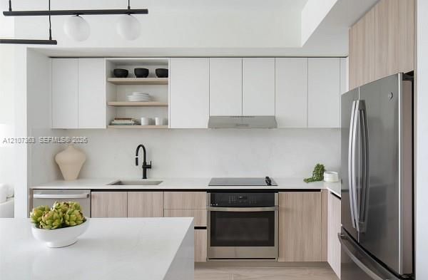 Kitchen featuring two-tone flat-panel cabinetry, light-toned stone-finish countertops, stainless steel appliances, matte black hardware, and linear pendant lighting