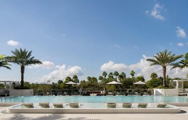 Swimming pool featuring a sun shelf with built-in lounge areas, integrated water fountains, and paved decking surrounded by mature palm trees