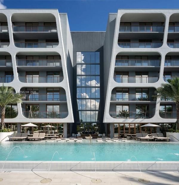 Contemporary architectural exterior featuring modular concrete framing, full-height glass curtain wall, integrated tiered balconies with glass railings, and a ground-level swimming pool with built-in water fountains