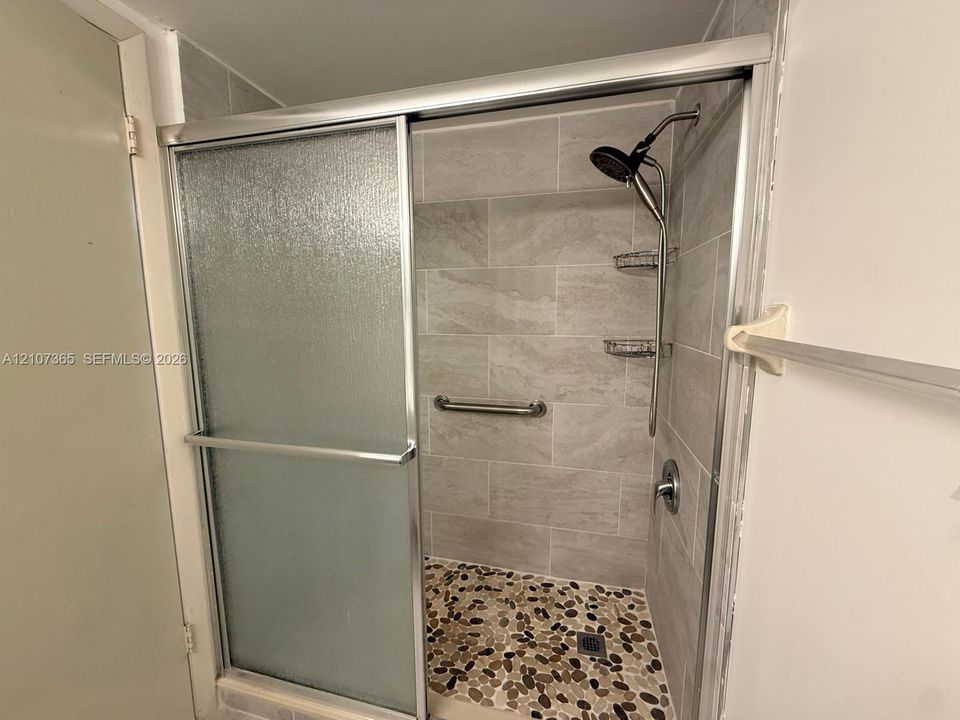 Walk-in shower featuring textured glass sliding doors, stone pebble floor tiling, large-format wall tile, a metal grab bar, and integrated storage shelving