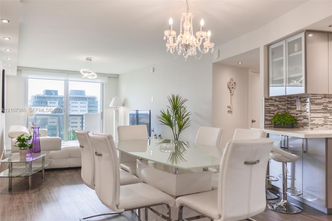Open-concept living space featuring wood-finish flooring, a multi-tiered crystal chandelier, recessed lighting, and a kitchen with glass-front cabinetry and stone-tiled backsplash