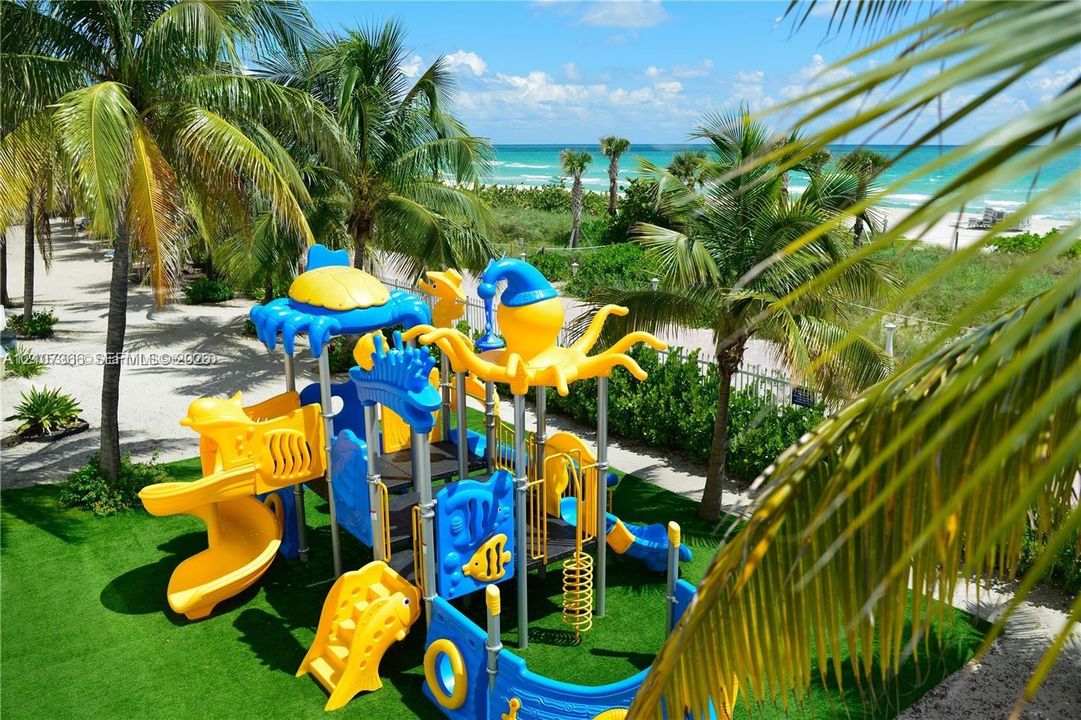 Outdoor play structure situated on artificial turf featuring mature palm trees and direct ocean proximity