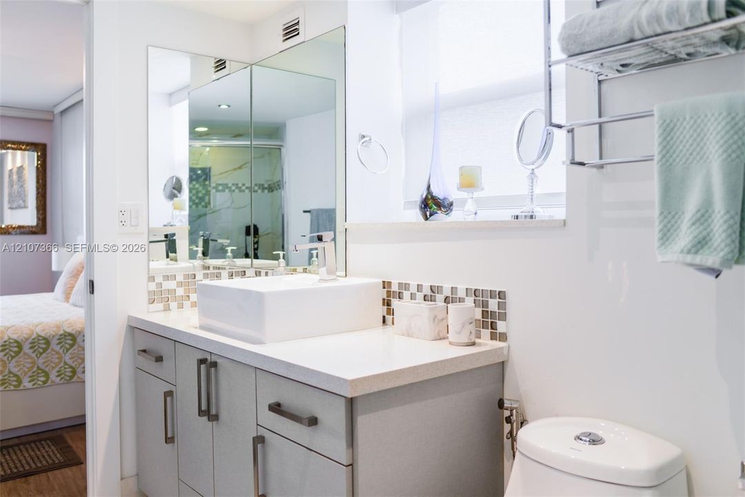 Bathroom vanity featuring a rectangular vessel sink, polished stone-surface countertop, mosaic tile backsplash, and light gray cabinetry with metallic hardware