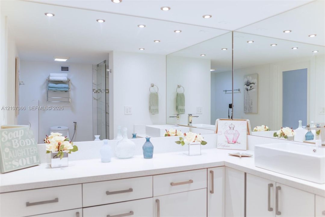 Bathroom featuring a white dual-sink vanity with matte metallic hardware, expansive wall-to-wall mirror, recessed ceiling lighting, and a glass-enclosed shower