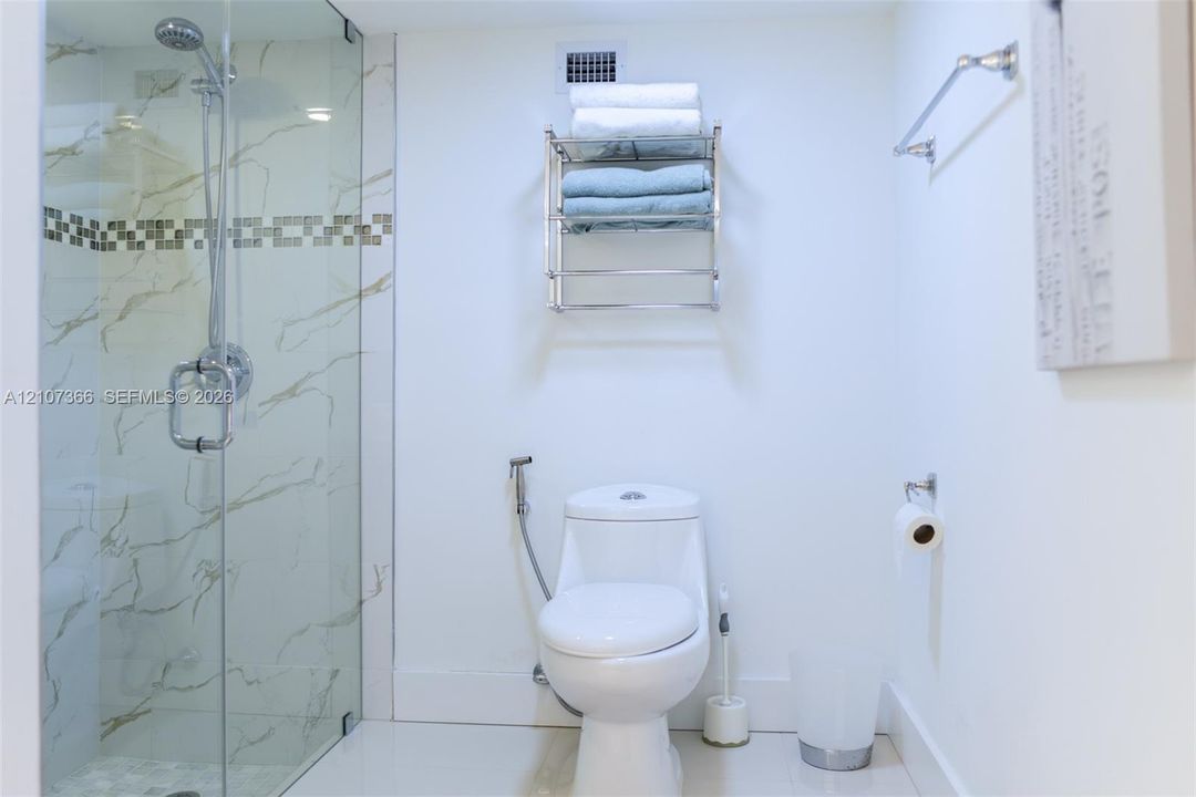 Walk-in glass-enclosed shower featuring large-format wall tiling with decorative mosaic accent, wall-mounted metal shelving unit, and bidet sprayer