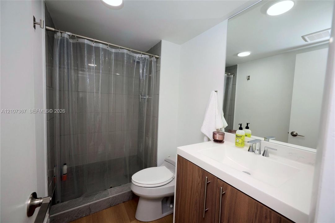 Bathroom featuring a vanity with a wood-finish cabinet and white integrated countertop, large wall-mounted mirror, recessed ceiling lighting, and a tiled shower surround