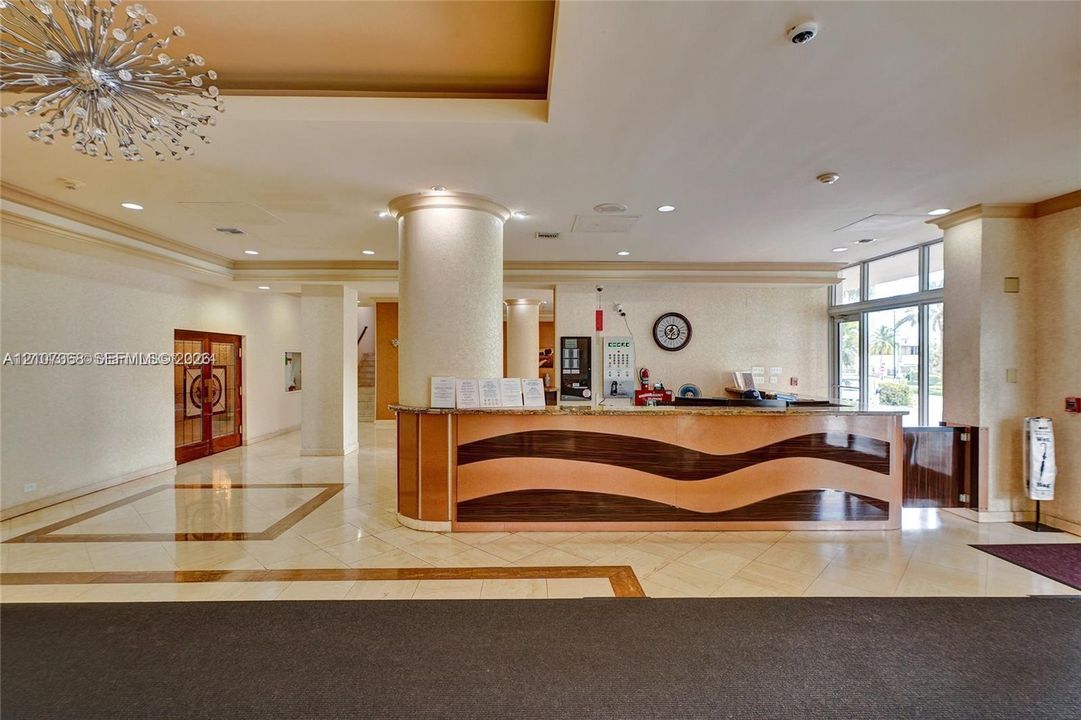 Lobby featuring a polished stone-tile floor with decorative inlay, a custom curved reception desk with wood-tone wave detailing, structural columns, and a large multi-point contemporary chandelier