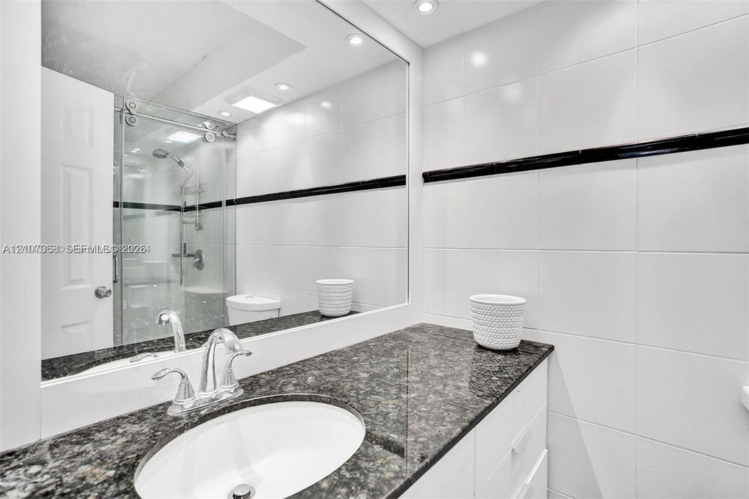 Bathroom featuring a vanity with stone-finish countertop, under-mount sink, polished chrome faucet, full-width mirror, and a glass-enclosed shower with white wall tiling accented by a horizontal dark tile border