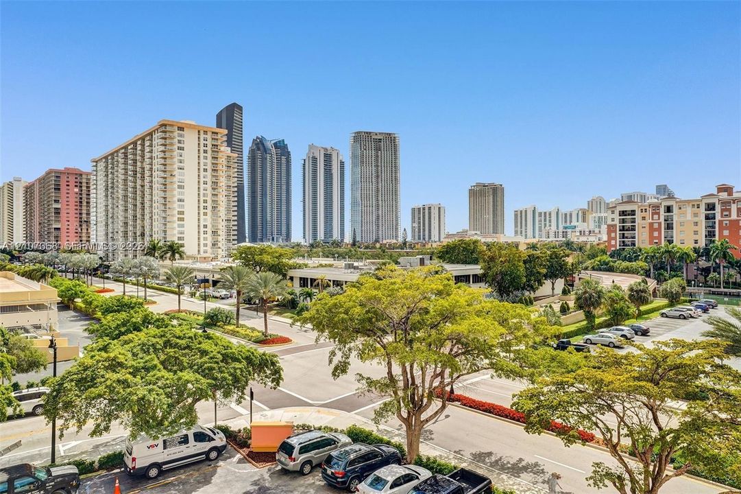 Urban skyline backdrop featuring high-rise buildings, mature deciduous trees, landscaped shrubbery, paved roadways, and surface parking areas