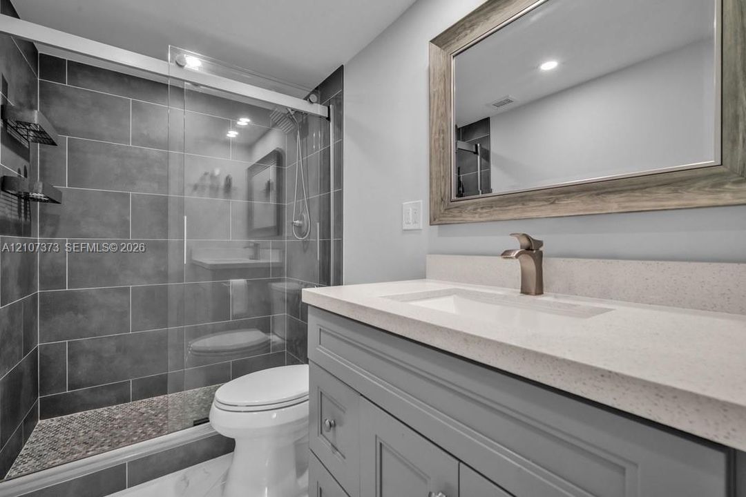 Bathroom featuring a vanity with a light-toned countertop, rectangular undermount sink, and brushed metal faucet, complemented by a walk-in shower with large-format grey tile surround and a glass sliding door