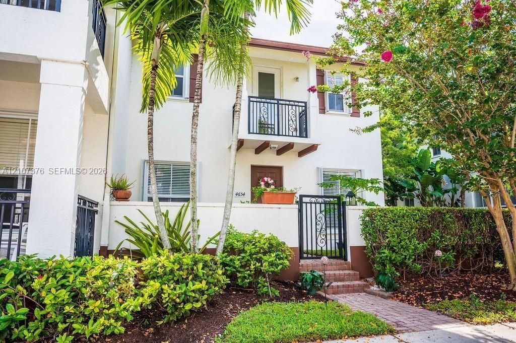 Stucco exterior featuring a second-story balcony with metal railing, a gated brick walkway entrance, and mature palm landscaping