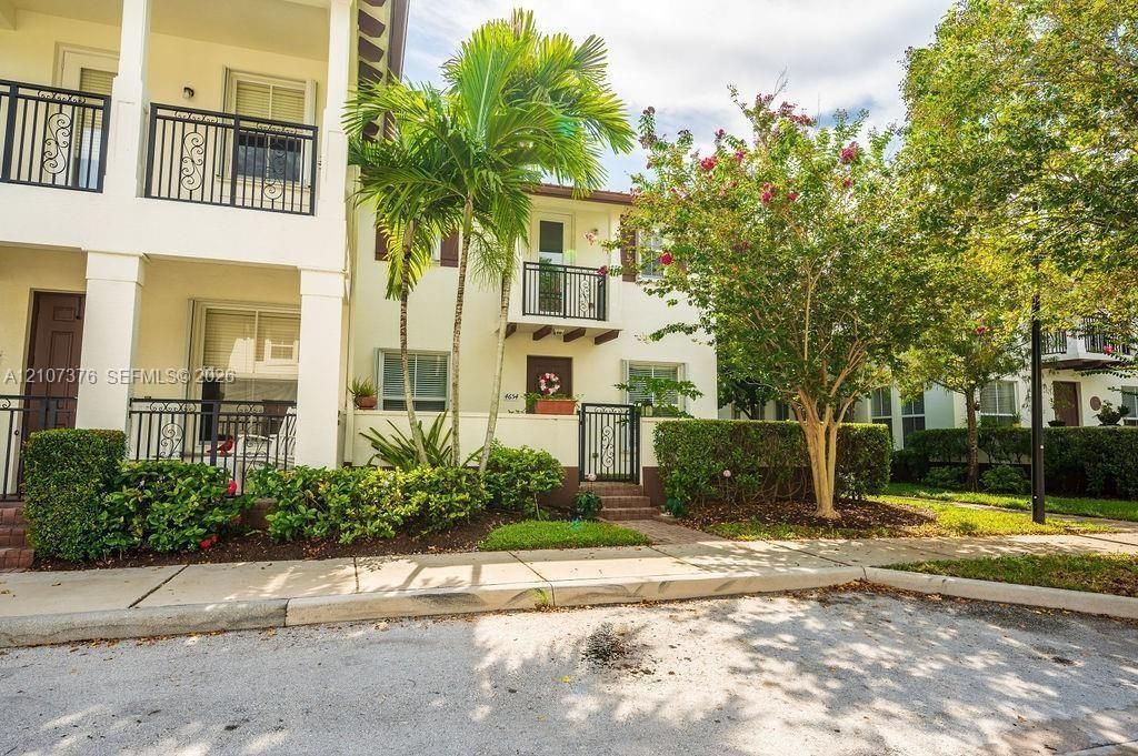 Multi-story exterior featuring light-toned stucco siding, decorative black metal railings, a gated entryway, and established tropical landscaping