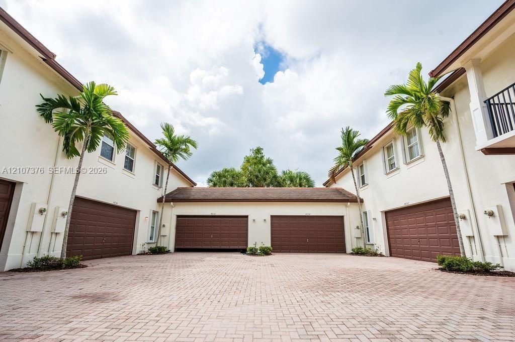 Stucco exterior featuring multiple garage bays, a herringbone-patterned paver driveway, and integrated palm tree landscaping
