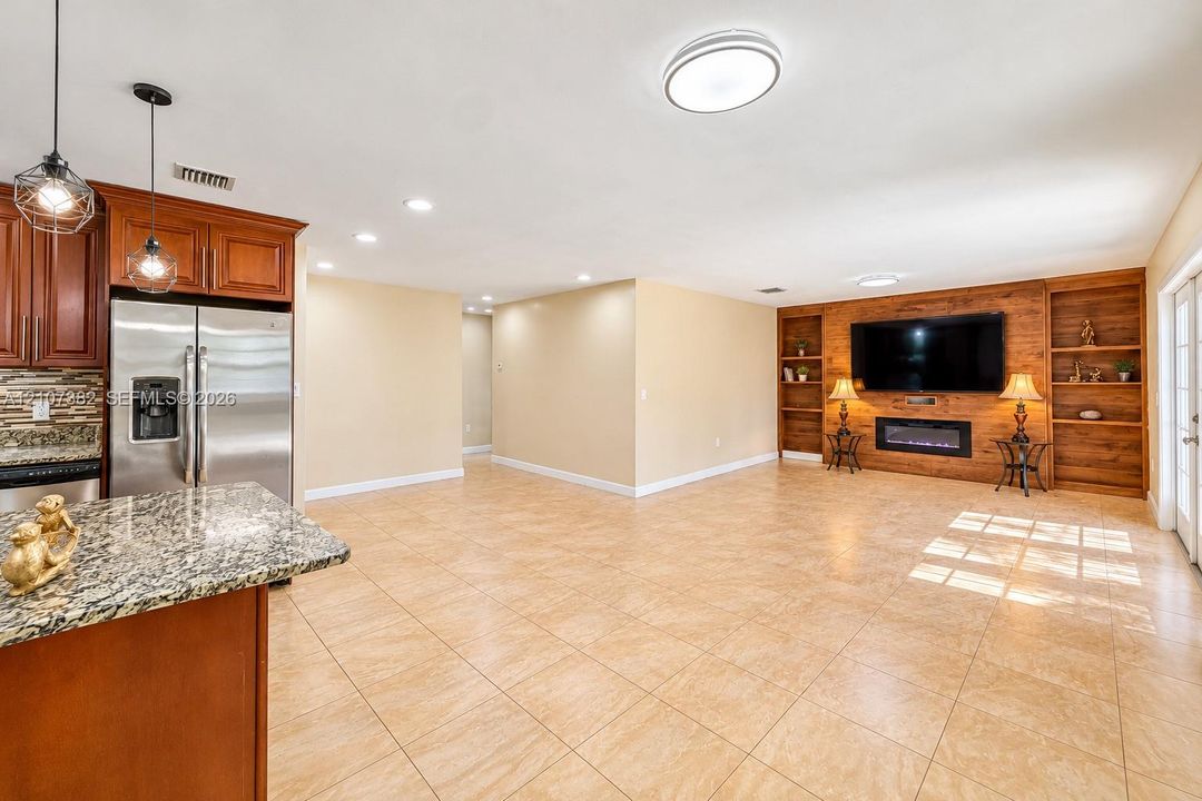 Open kitchen featuring stone countertops, custom wood cabinetry, and stainless steel appliances, anchored by a living area with a wood-paneled accent wall, built-in shelving, and an electric fireplace