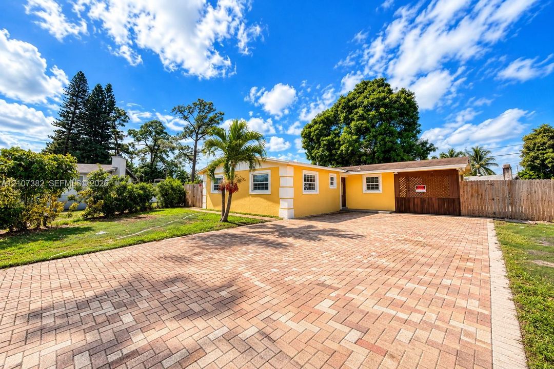 Exterior facade featuring a yellow stucco finish, expansive brick-paver driveway, integrated carport, and wood-latched privacy fencing