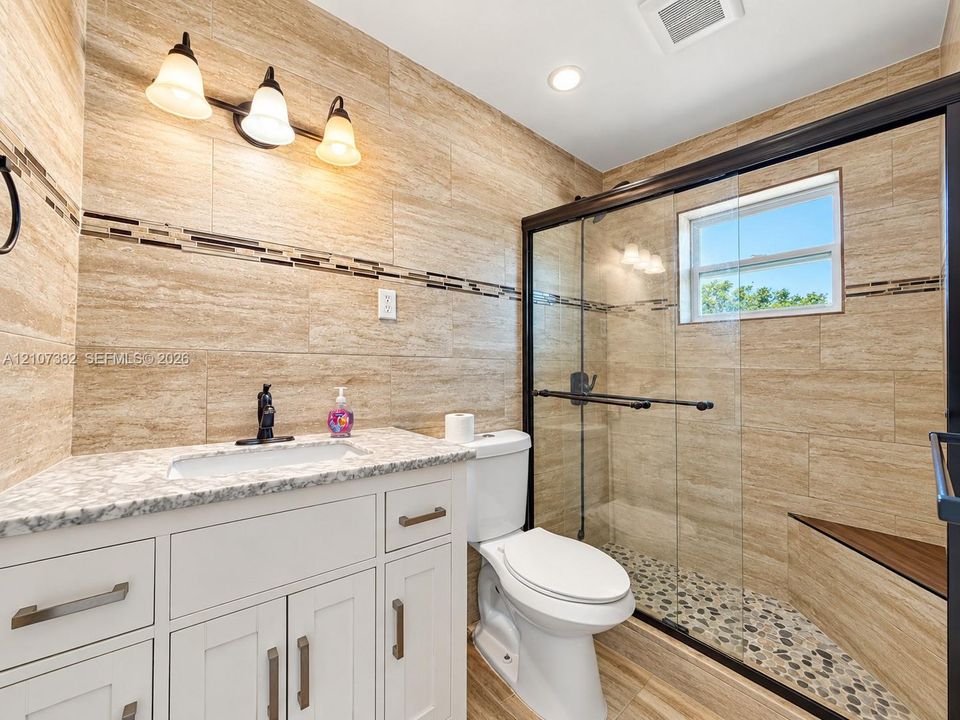 Bathroom featuring large-format wall tiling with decorative mosaic inlay, stone-composite countertop with integrated sink, shaker-style cabinetry, and a walk-in shower with pebble-stone flooring and a built-in bench