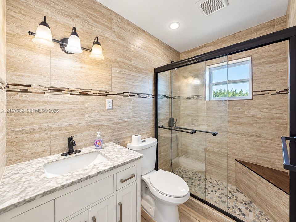 Full bathroom featuring floor-to-ceiling stone-look wall tile, vanity with a patterned countertop, glass-enclosed shower with pebble-stone flooring, and a built-in shower bench
