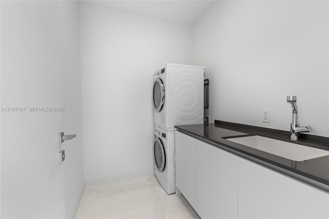 Laundry room featuring stacked washer and dryer, flat-panel cabinetry, dark stone-finish countertop, undermount sink, and high-arc chrome faucet