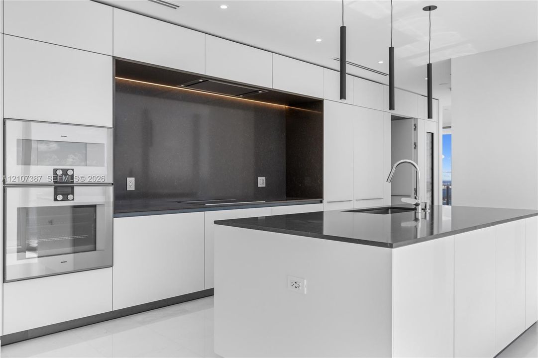Contemporary kitchen featuring floor-to-ceiling flat-panel cabinetry, a central island with a solid-surface countertop, integrated wall-mounted ovens, a dark stone-finish backsplash, and minimalist pendant lighting