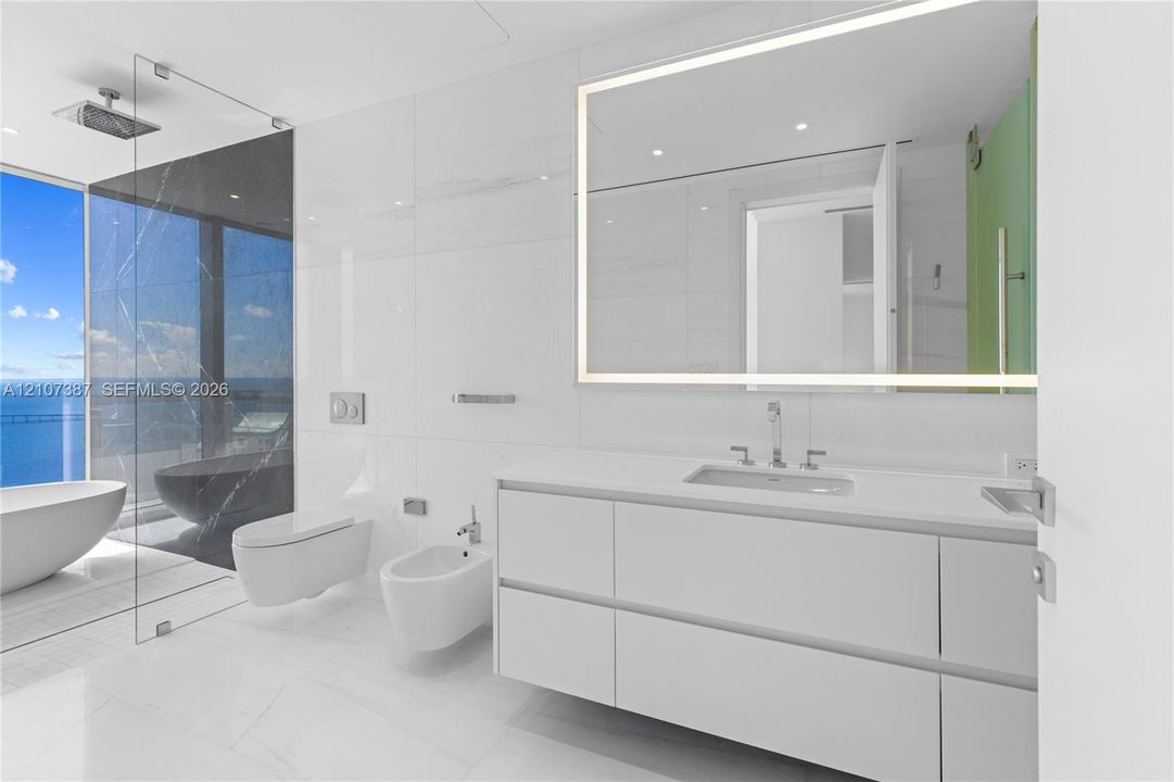 Modern floating vanity with a white countertop and rectangular sink, LED-backlit mirror, wall-mounted commode and bidet, and floor-to-ceiling glass-enclosed shower area