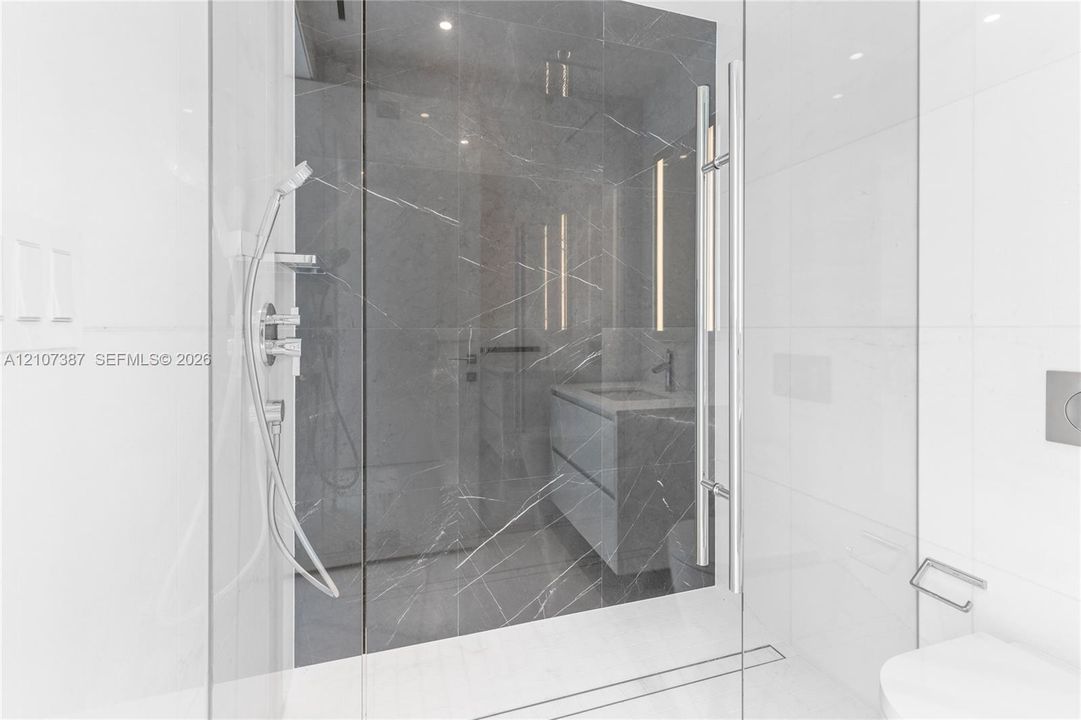 Walk-in shower featuring large-format dark veined tile surround, frameless glass enclosure, handheld shower fixture, and linear drain