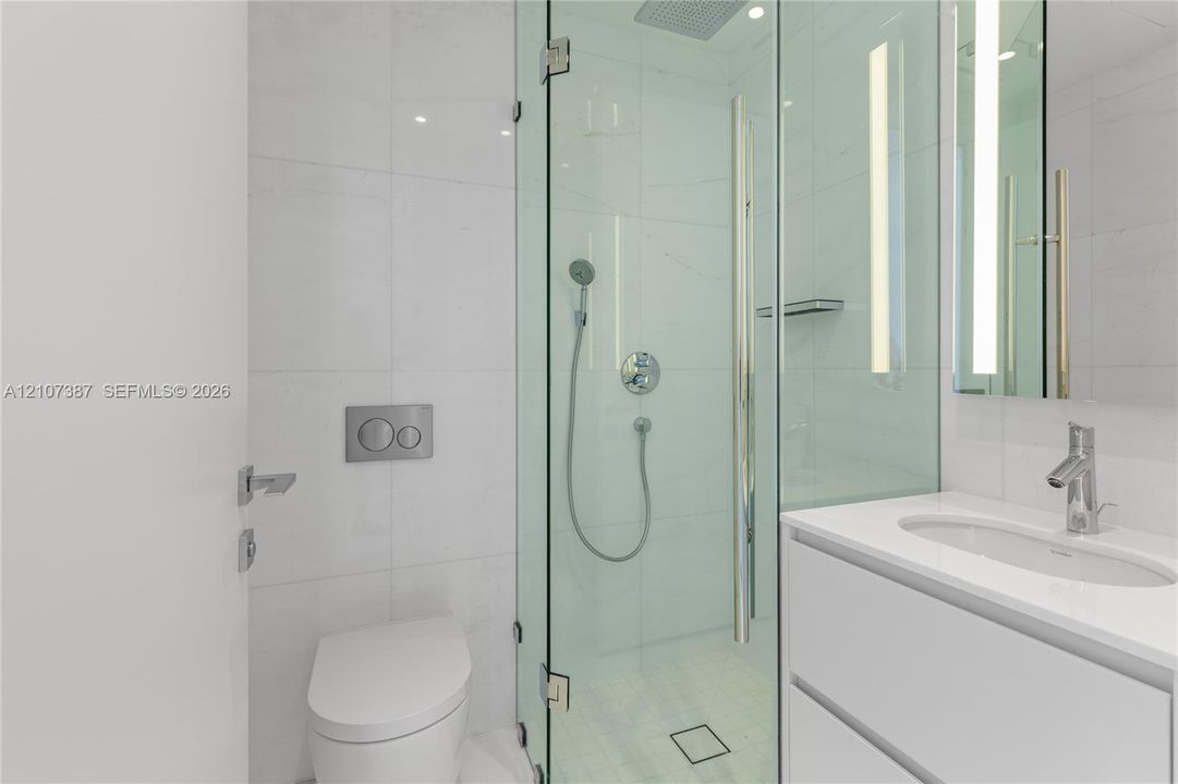 Bathroom featuring a glass-enclosed shower with a rainfall showerhead, wall-mounted dual-flush toilet, white vanity with an integrated sink, polished chrome fixtures, and large-format tile surround
