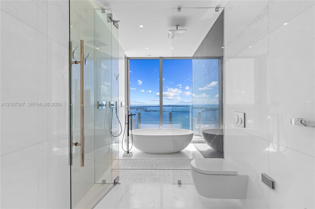 Freestanding soaking tub, floor-to-ceiling glass-enclosed shower, wall-mounted commode, large-format tile flooring, and floor-to-ceiling windows with coastal scenery