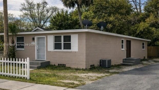 Single-story masonry exterior featuring white louvered shutters, a white picket fence, a central metal security entry door, and an asphalt driveway