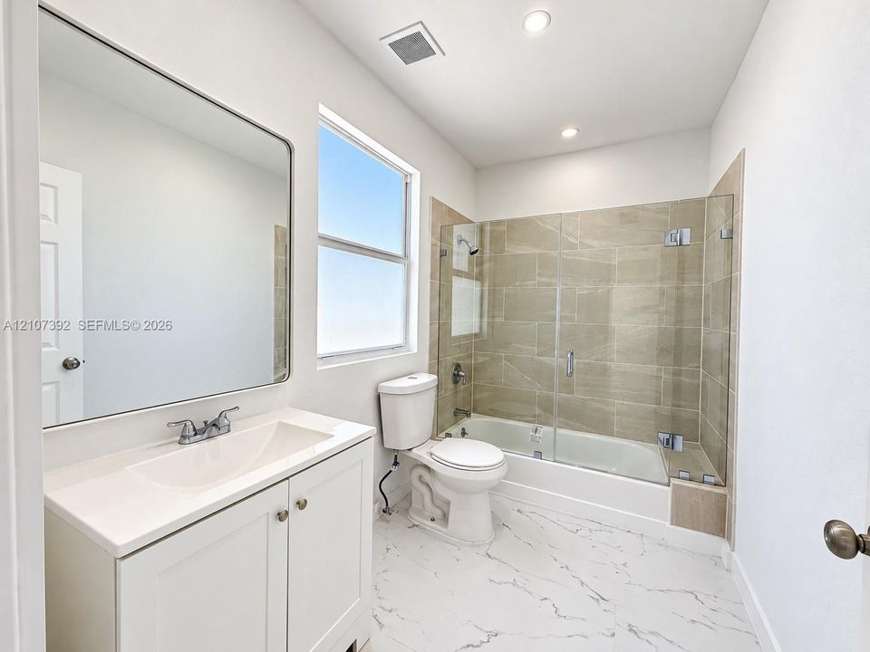 White cabinetry with a solid surface countertop, large rectangular mirror, tile-surround shower and tub combination with a glass panel, and large-format flooring