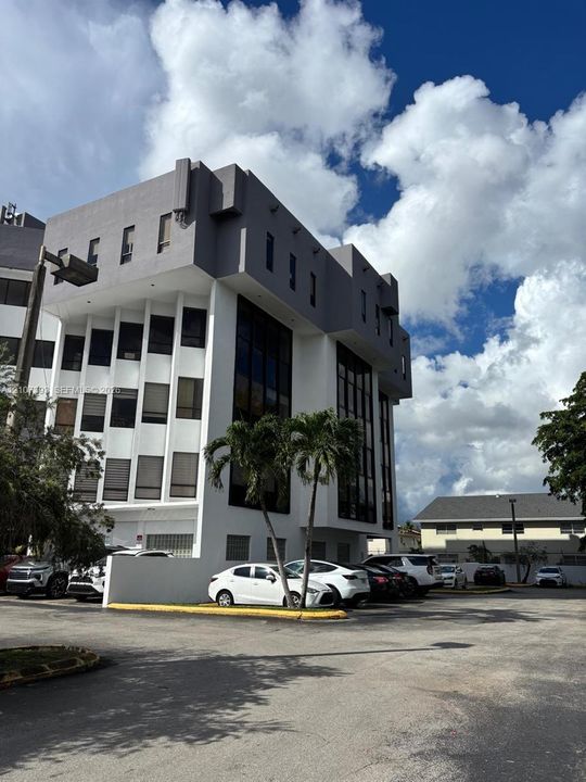 Multi-story exterior featuring a gray and white facade, glass curtain walls, vertical architectural columns, and surface-level parking