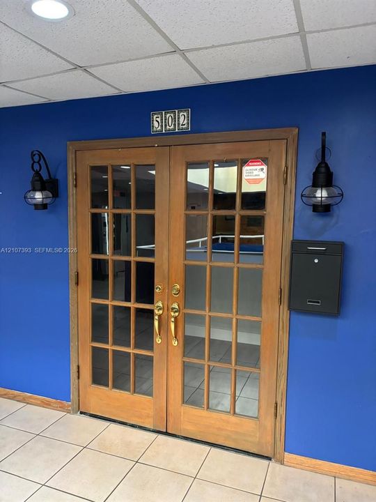 Double entry doors featuring a wood frame, glass pane detailing, and brass hardware, complemented by twin black wall-mounted lanterns and a modern wall-mounted metal mailbox