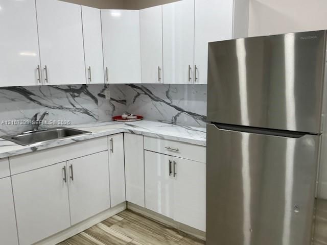 Kitchen featuring high-gloss white cabinetry, stone-patterned backsplash and countertops, stainless steel undermount sink with gooseneck faucet, full-size stainless steel refrigerator, and wood-finish flooring