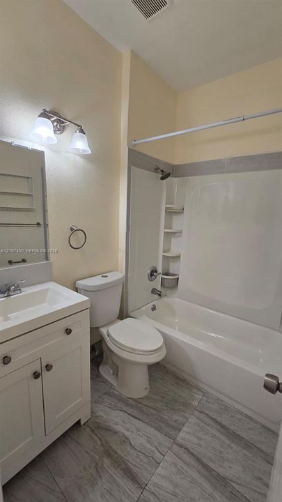 White vanity with rectangular integrated sink, neutral stone-finish flooring, two-light wall-mounted fixture, and combination tub and shower with built-in shelving