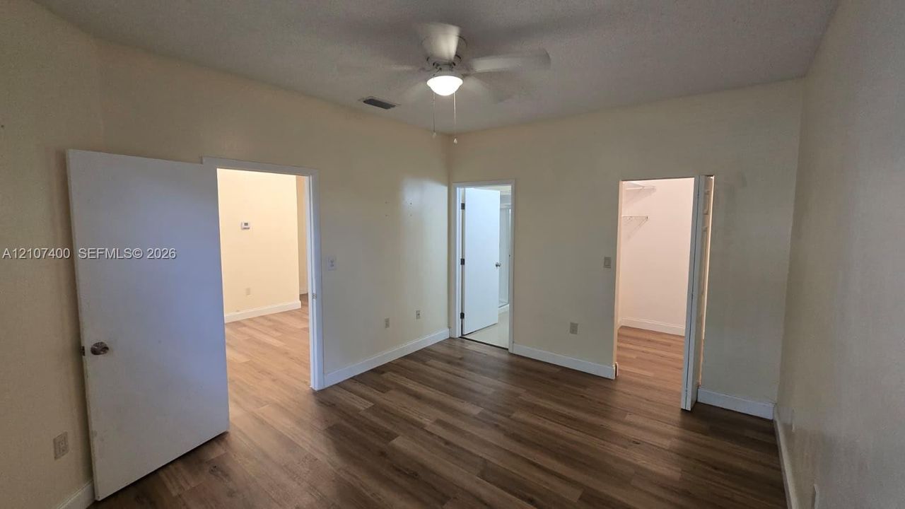 Room featuring wood-finish flooring, a central ceiling fan with integrated lighting, and multiple interior doorways leading to separate closet and storage areas