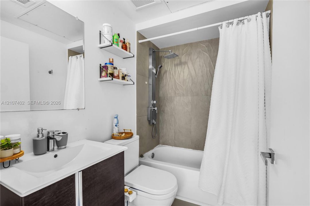 Bathroom featuring a dark wood-finish vanity with a white integrated basin, frameless wall-mounted mirror, two built-in wall shelves, a bathtub with textured tile surround, and a stainless steel shower panel system