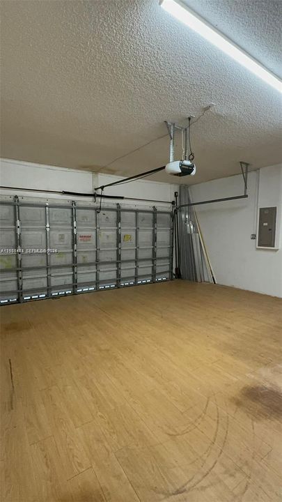 Enclosed garage featuring wood-finish flooring, metal sectional overhead door, ceiling-mounted automatic door opener, and integrated electrical service panel