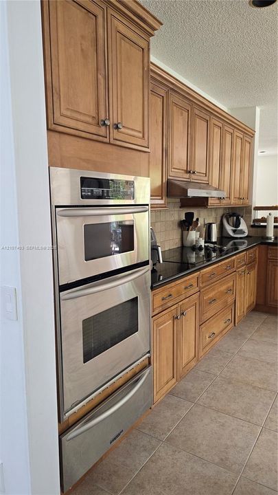 Kitchen featuring natural-finish wood cabinetry, dark stone countertops, tile backsplash, and built-in stainless steel double wall ovens