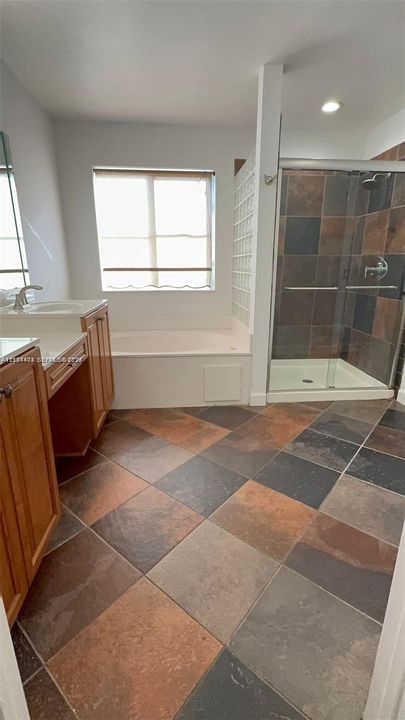 Bathroom featuring wood-finish cabinetry, a white countertop with an integrated sink, a deep soaking tub, a multi-tone tile floor, and a walk-in shower with glass doors